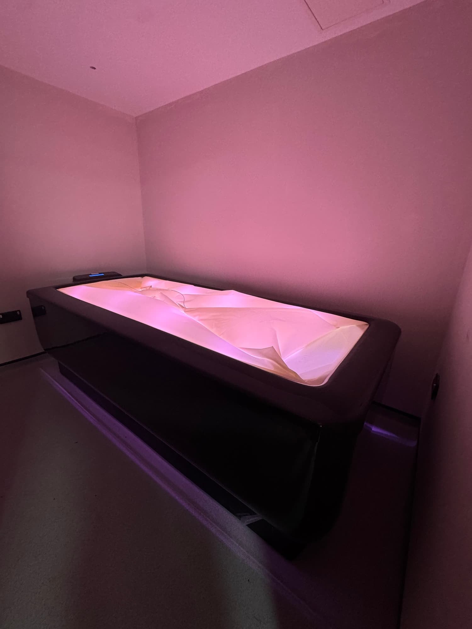 A modern sensory or relaxation bed with a soft pink glow, positioned in a corner against plain light-colored walls, in a room with minimal decor.