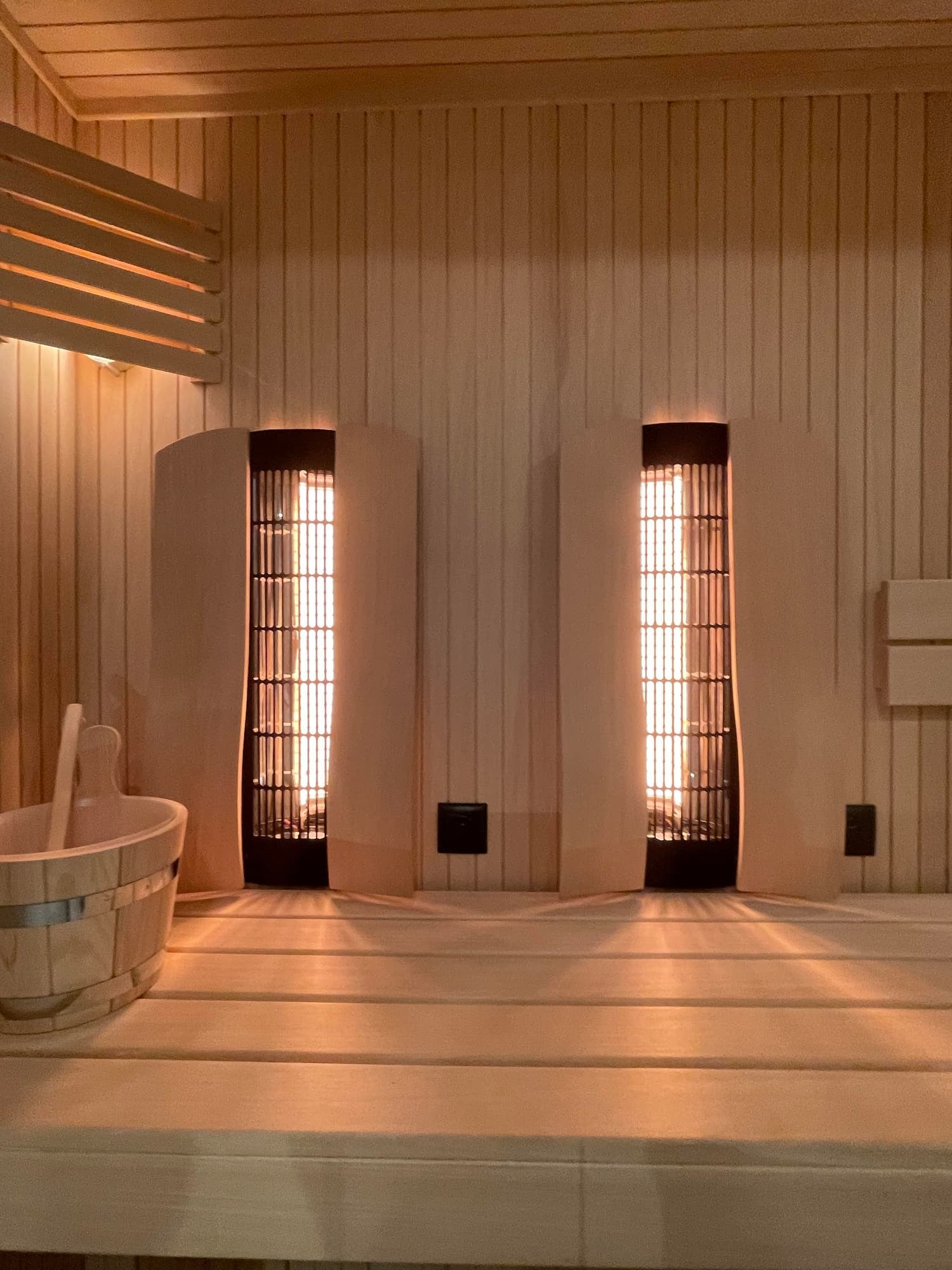 Two Infrared Saunas inside a wooden room with a wooden bench in the foreground